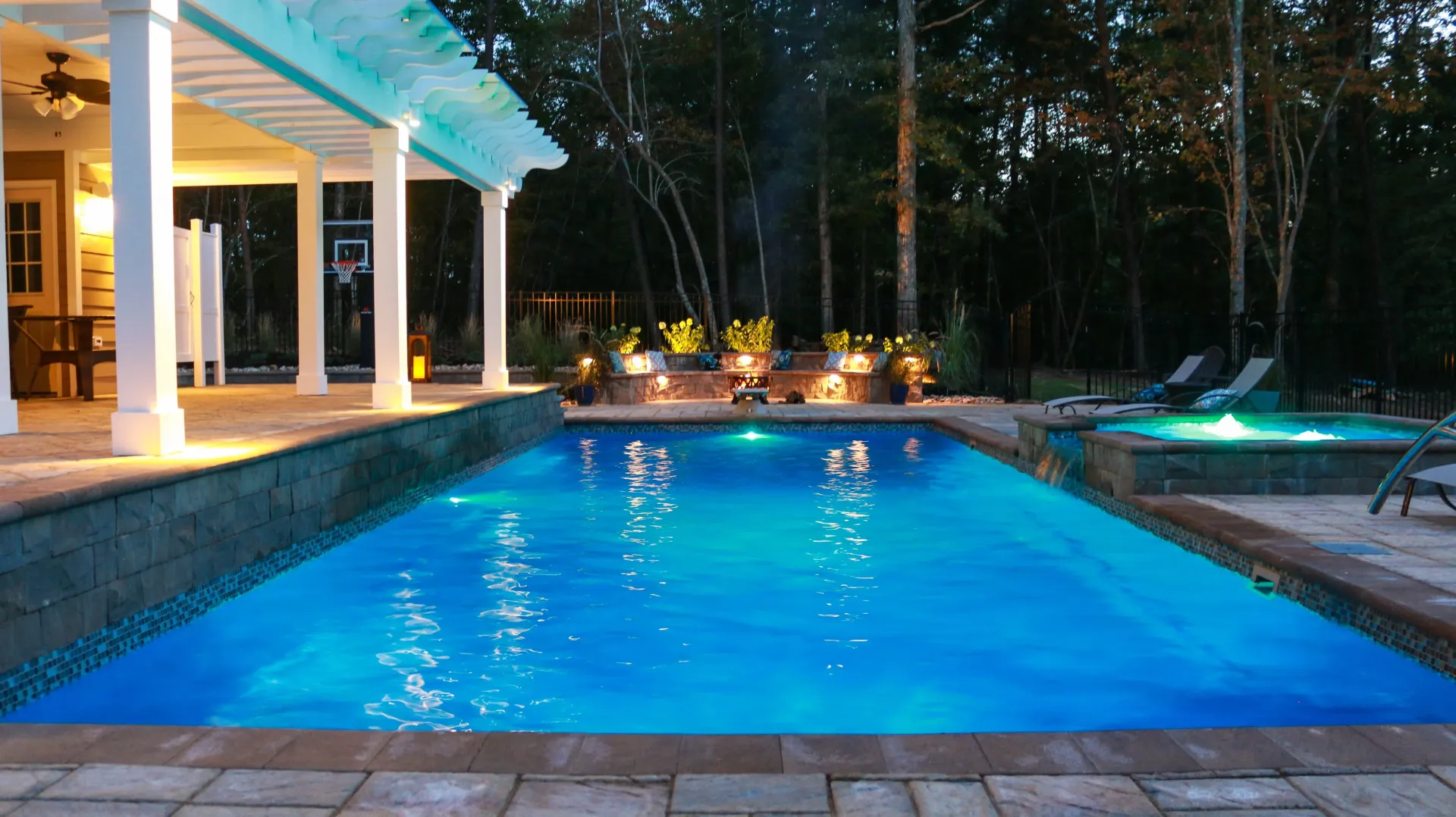 Blue-lit backyard pool at night beside a lit patio and trees