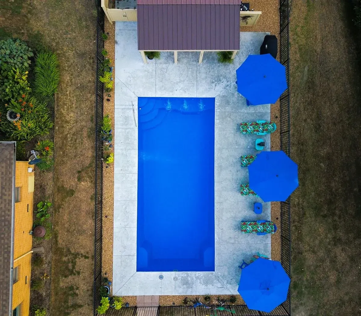 Aerial view of a rectangular blue swimming pool with three blue umbrellas and lounge chairs on a patio.