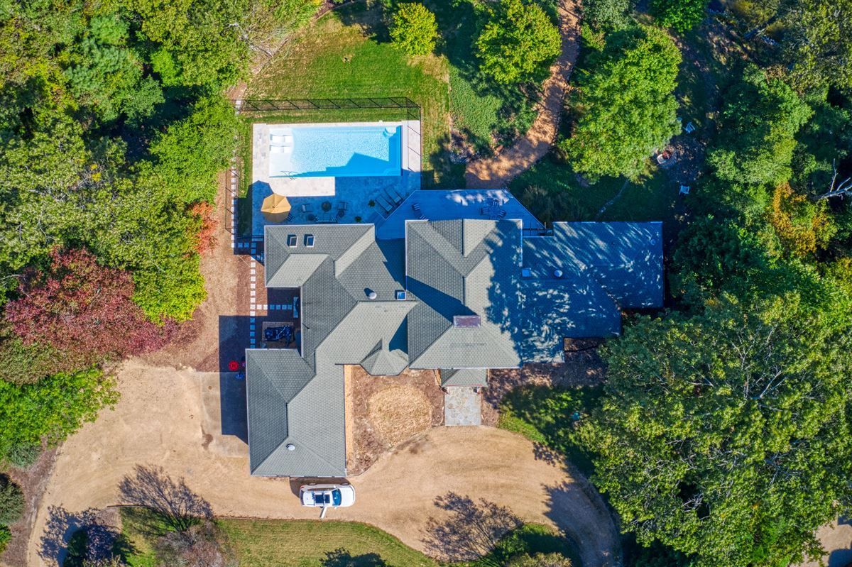 Aerial view of a house with a blue pool, surrounded by dense green trees and a driveway.