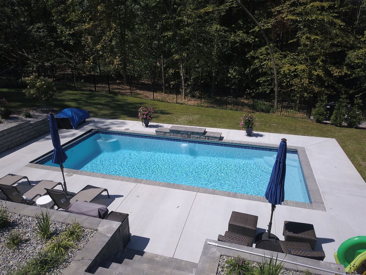 Rectangular backyard pool with blue water, lounge chairs, umbrellas, and trees surrounding the deck.