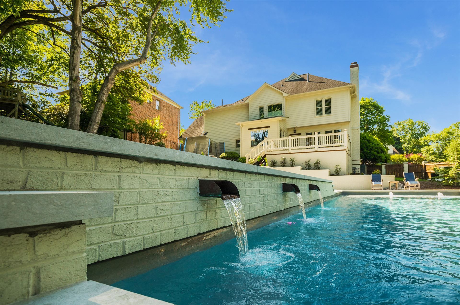 Outdoor pool with a stone water spout, cream-colored house, and trees in bright sunlight