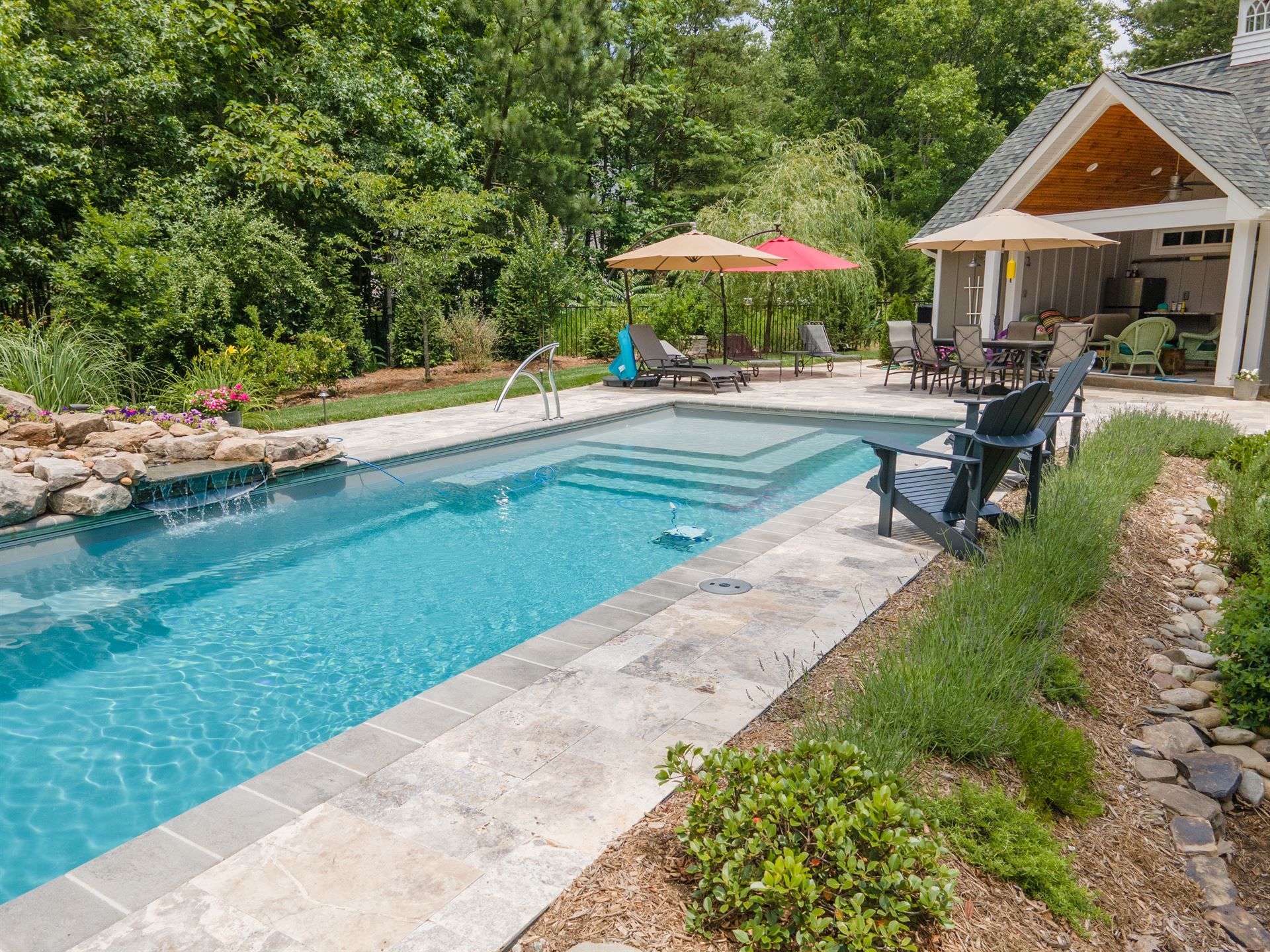Backyard pool beside a patio and cabana, with lounge chairs, umbrella, and lush greenery.