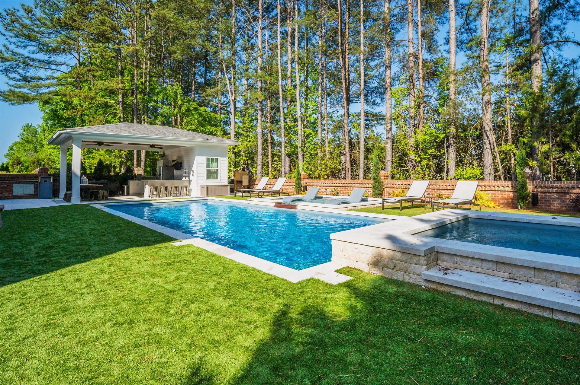 Backyard pool with blue water, white cabana, green lawn, and tall trees in the background