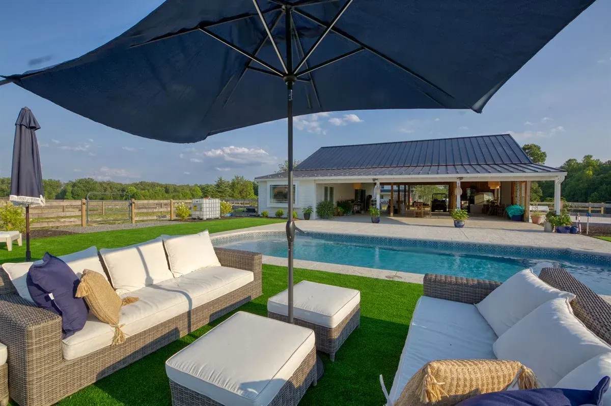 Modern patio with white seating, blue umbrella, and poolside view of a house under a clear sky