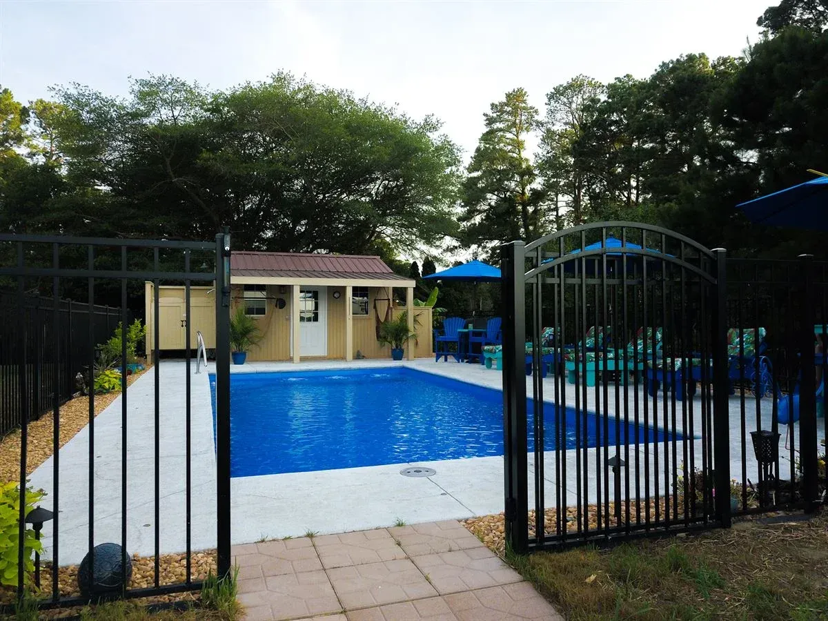 Backyard swimming pool behind black metal gate, with small building and trees in the background