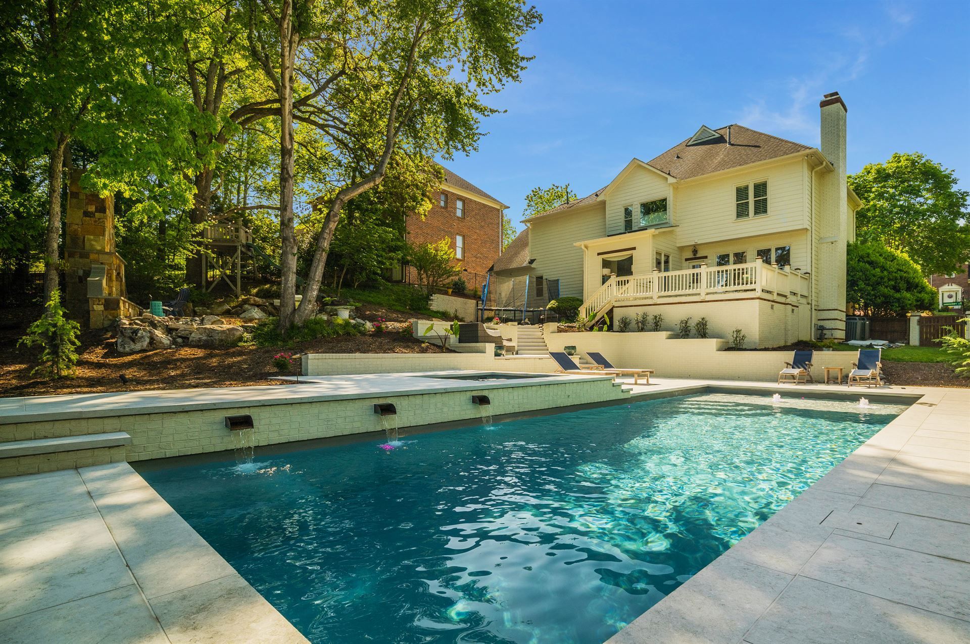 Backyard pool beside a cream-colored house, surrounded by tall trees and a sunlit patio.