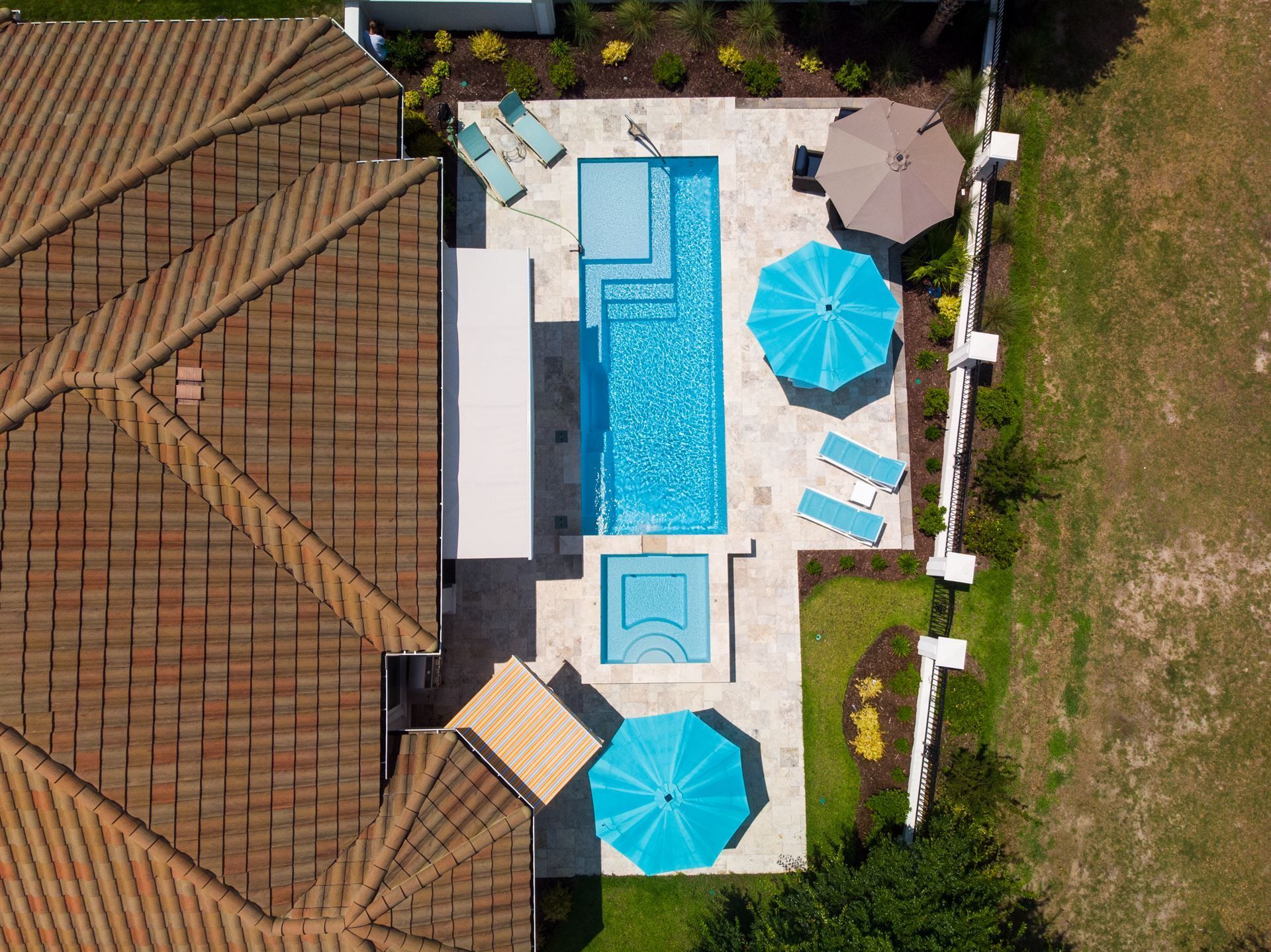 Aerial view of a backyard pool with blue umbrellas, loungers, and patio beside a tiled roof and lawn