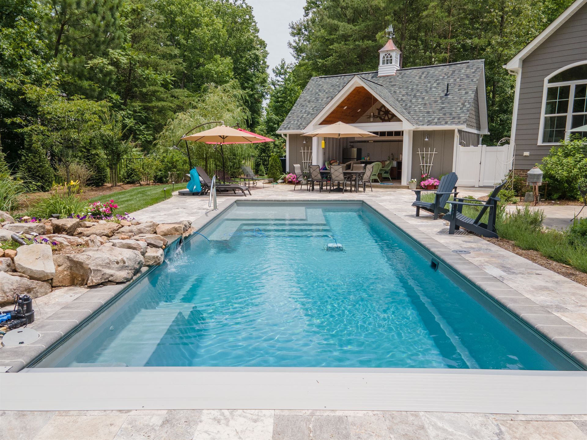 Backyard swimming pool beside a stone patio and pool house, surrounded by trees and lounge chairs