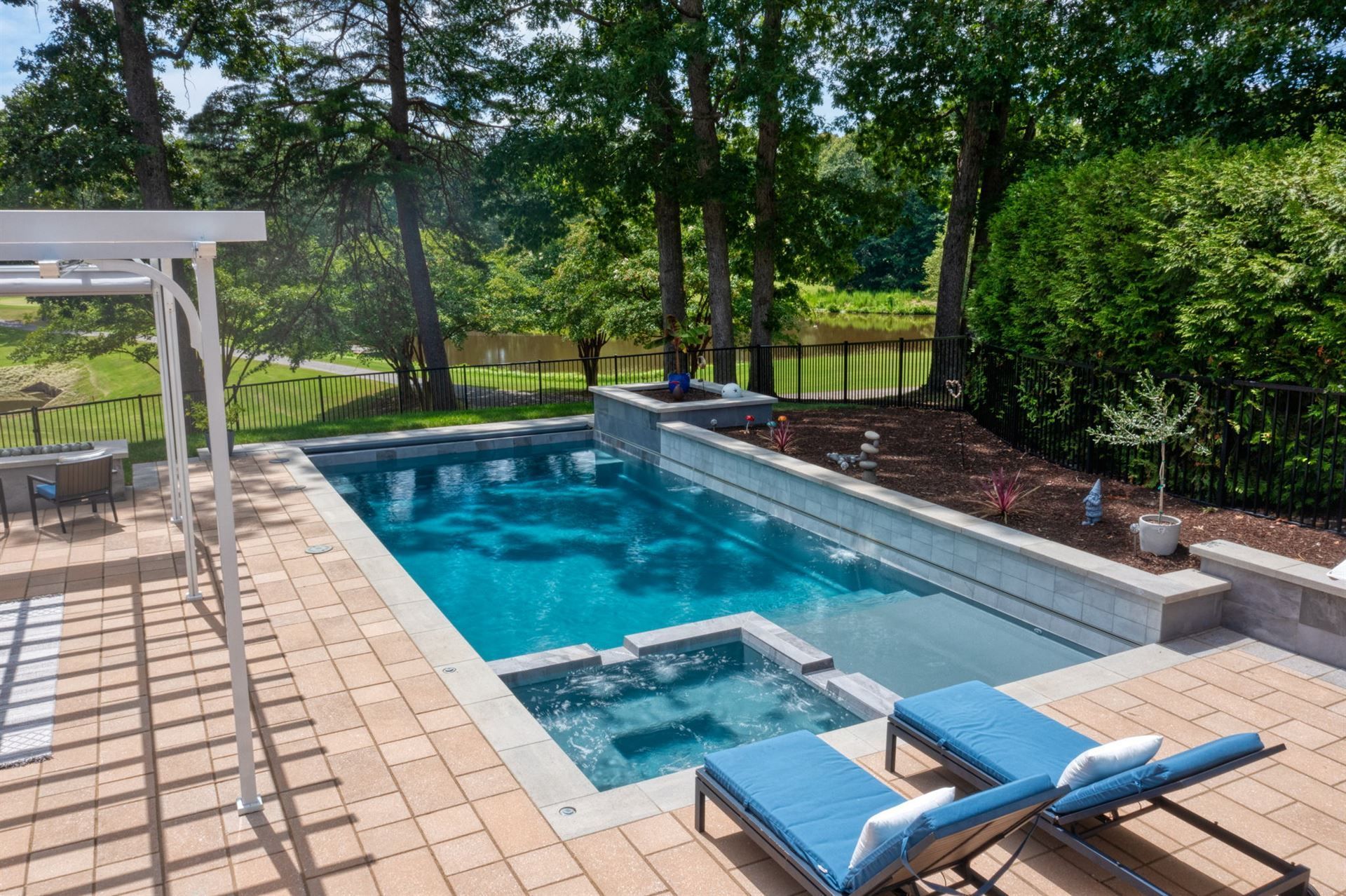 Backyard pool with hot tub, blue lounge chairs, and green trees beyond the patio