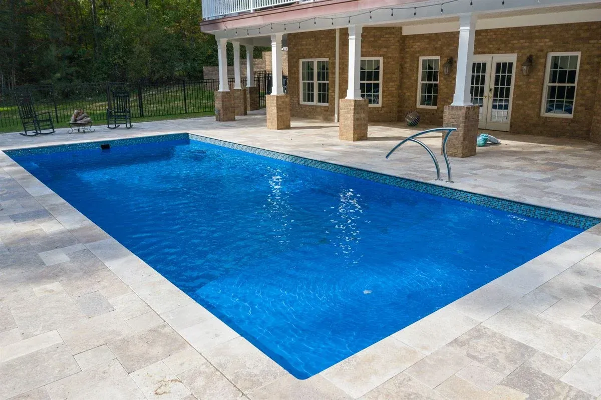 Rectangular blue swimming pool beside a stone patio and house with a metal handrail