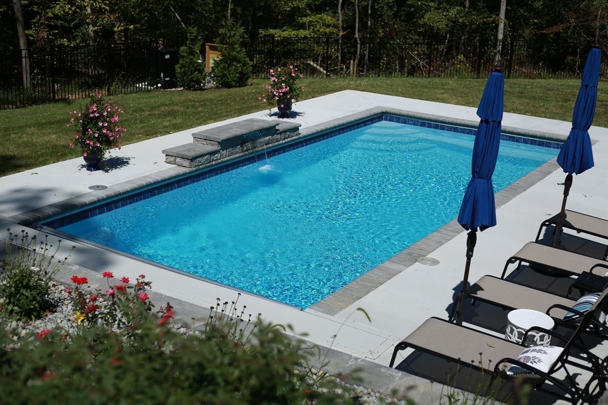 Rectangular outdoor swimming pool with blue water, lounge chairs, and closed blue umbrellas on a sunny patio.