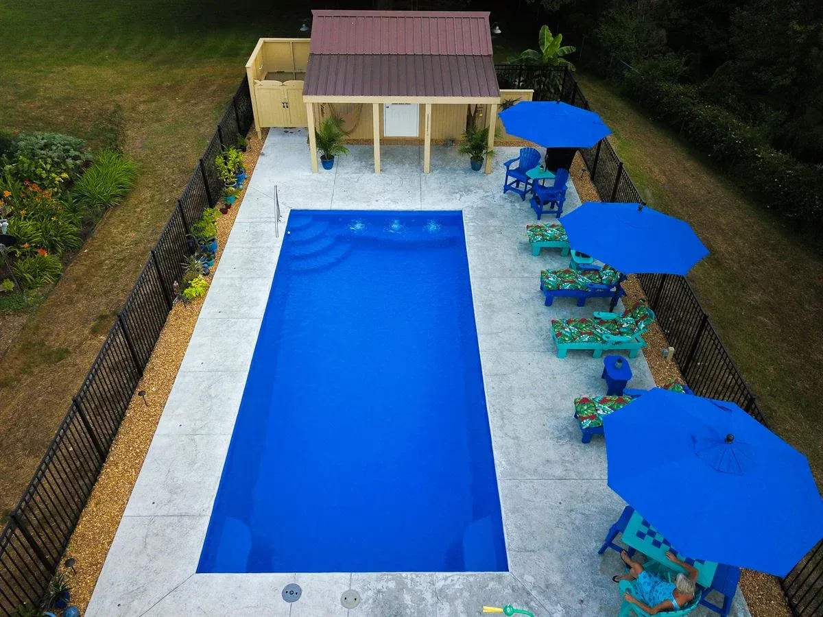 Backyard swimming pool with blue umbrellas and lounge chairs beside a small pool house