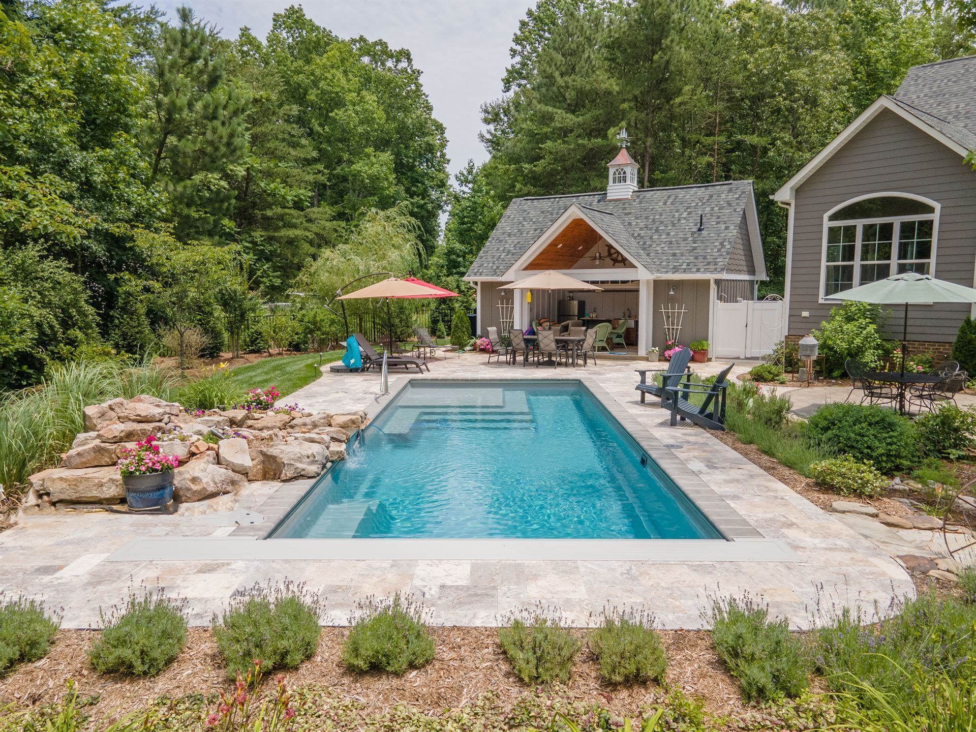 Backyard pool with stone patio, lounge chairs, and a small house surrounded by trees