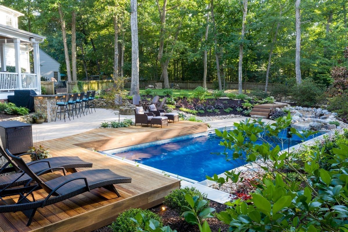 Backyard pool with lounge chairs, patio seating, and trees in a wooded setting