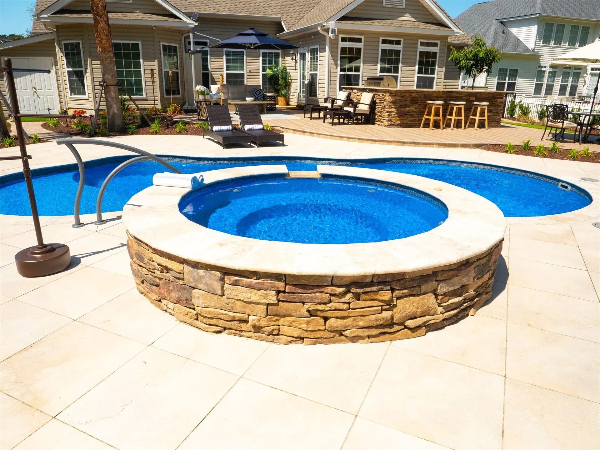 Backyard patio with circular stone hot tub, blue pool, and lounge chairs near a house