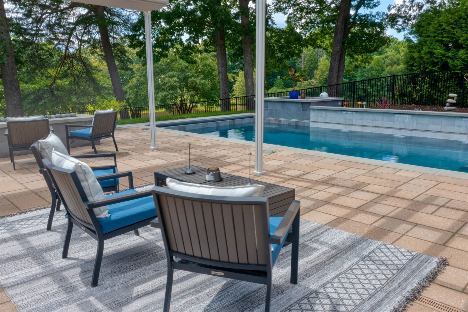 Patio seating beside a backyard swimming pool with blue cushions and a gray rug under trees