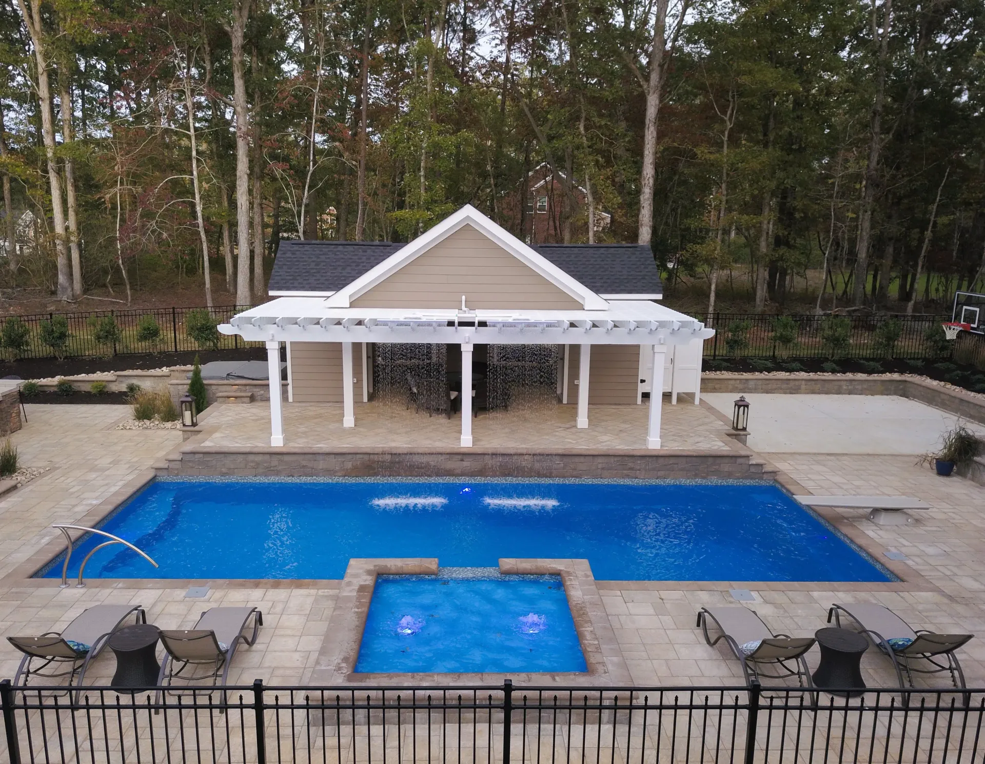 Backyard pool with a small spa, white pavilion, lounge chairs, and wooded backdrop