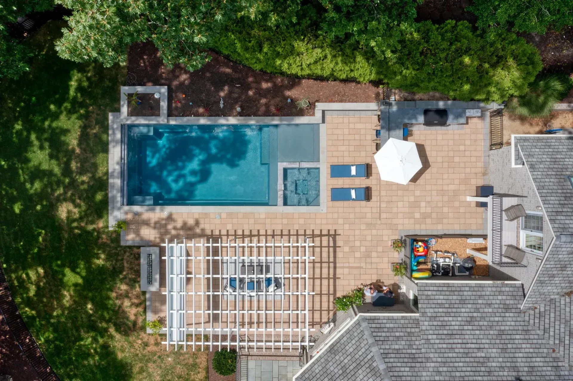 Aerial view of a backyard pool, patio deck, lounge chairs, and outdoor seating surrounded by trees