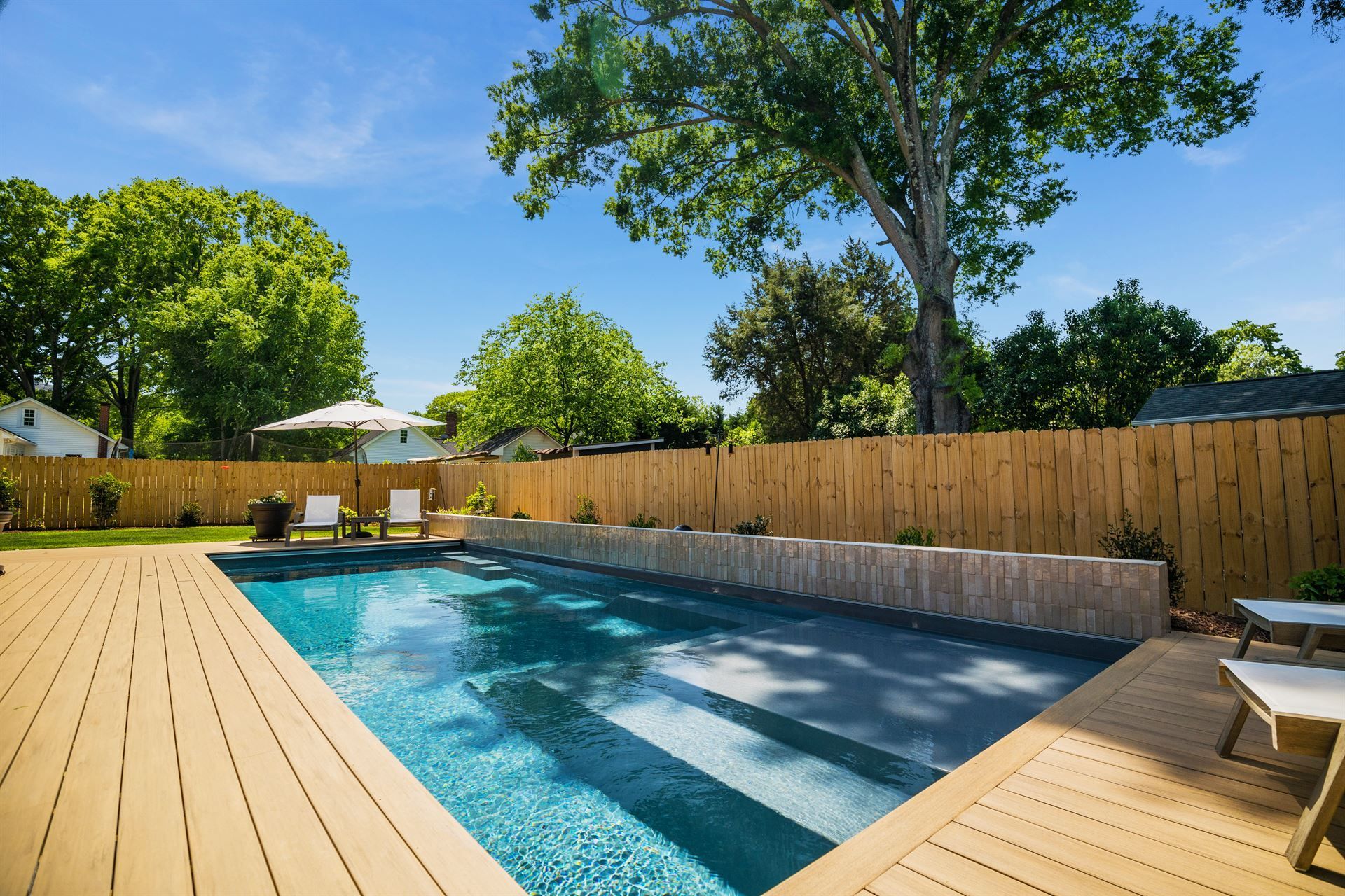 Backyard swimming pool with wooden deck, lounge chairs, and tall trees under a clear blue sky
