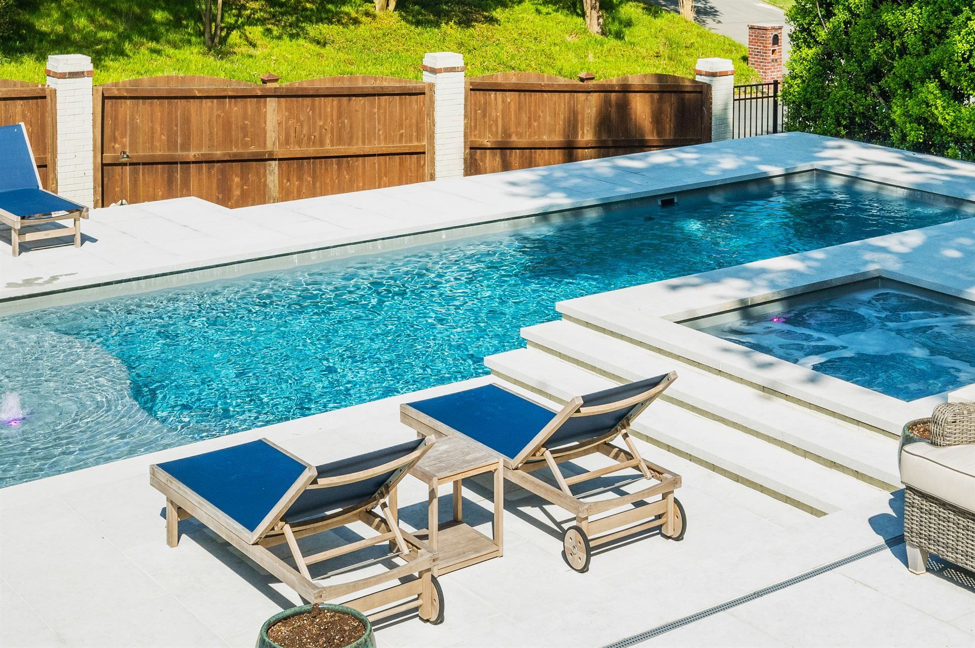 Modern backyard pool with blue water, white deck, lounge chairs, and a hot tub beside a wooden fence