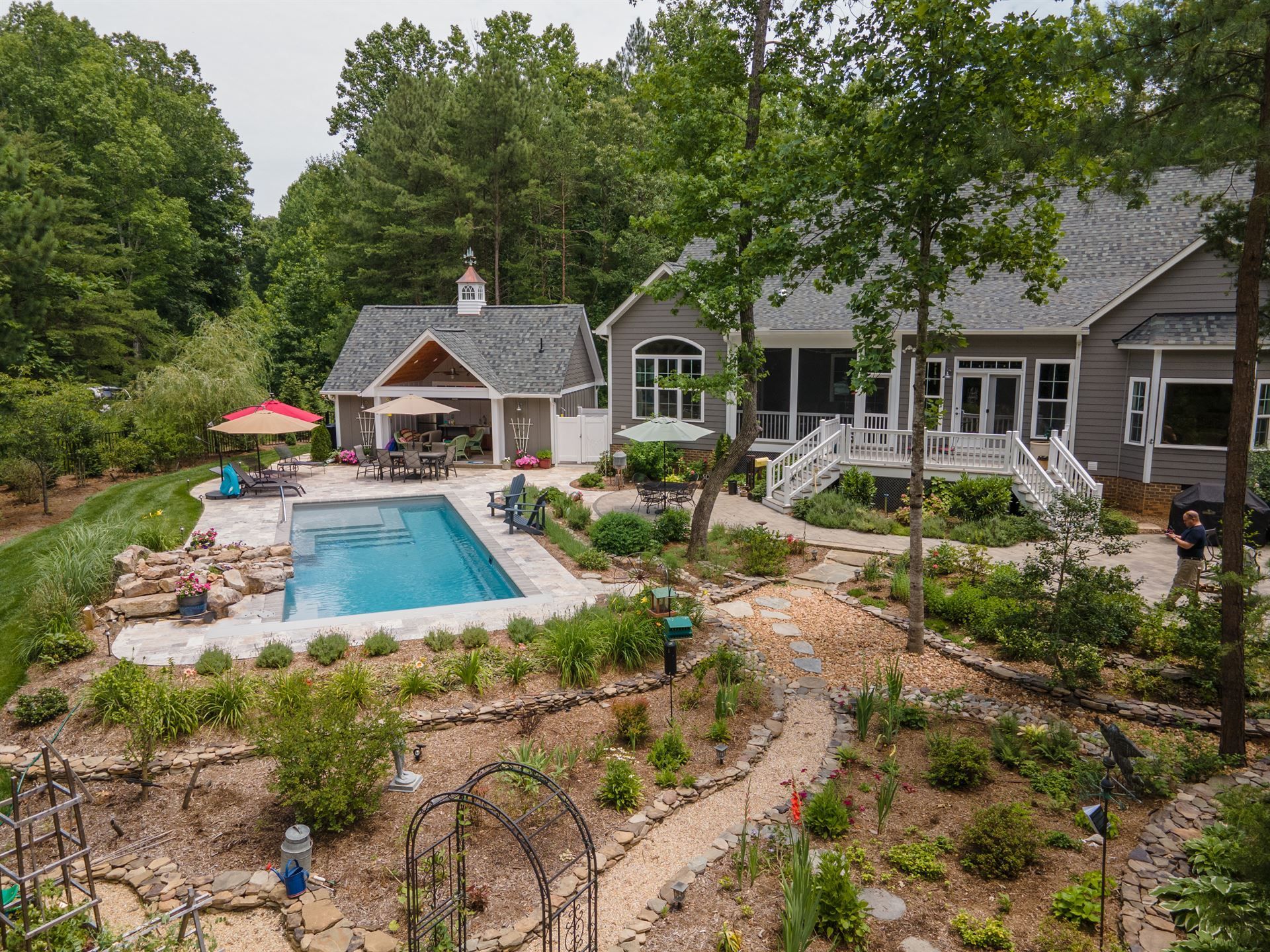 Backyard with a pool, stone patio, umbrella, and gray house surrounded by trees and landscaping