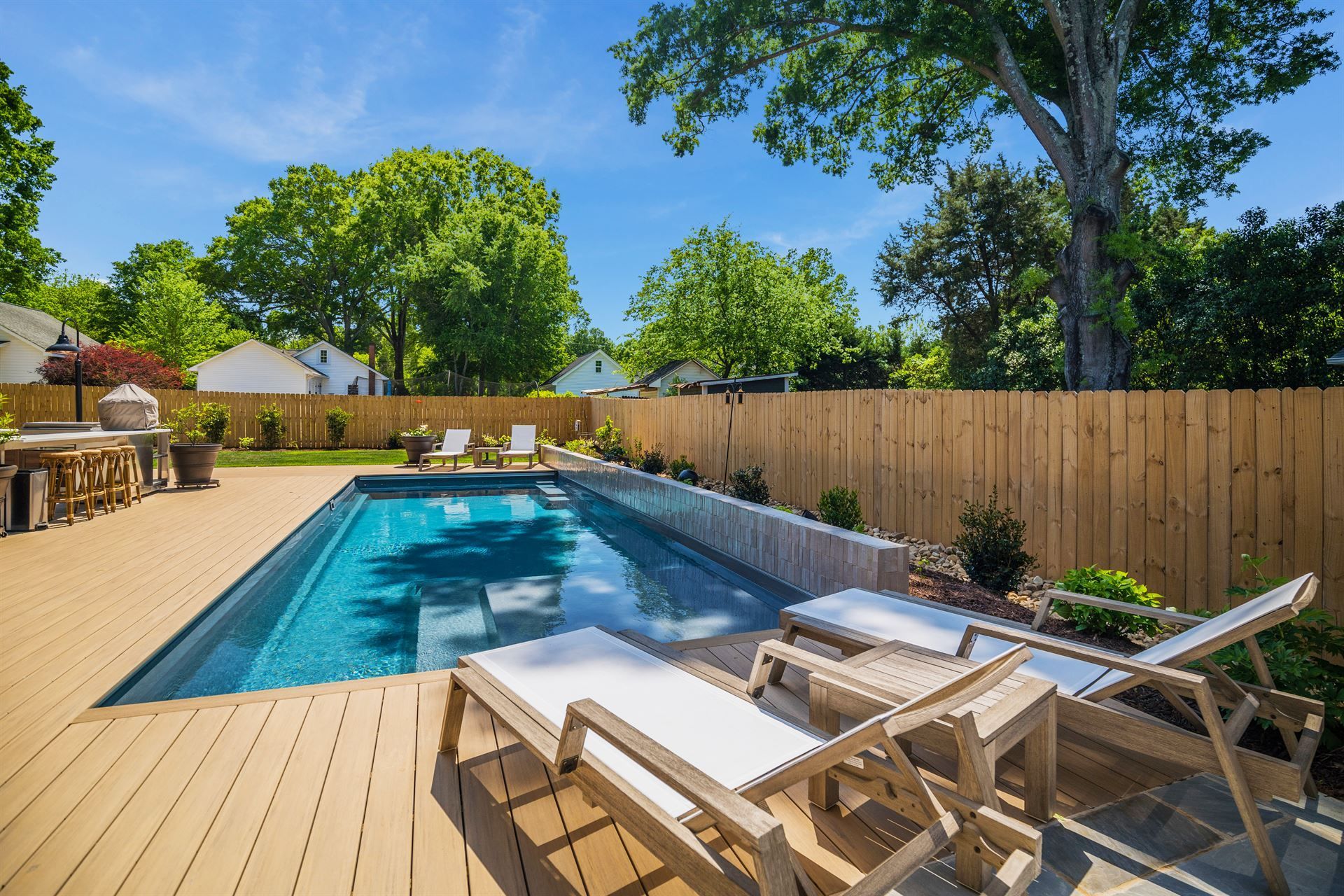 Backyard patio with a narrow blue pool, lounge chairs, wooden deck, and tall trees on a sunny day