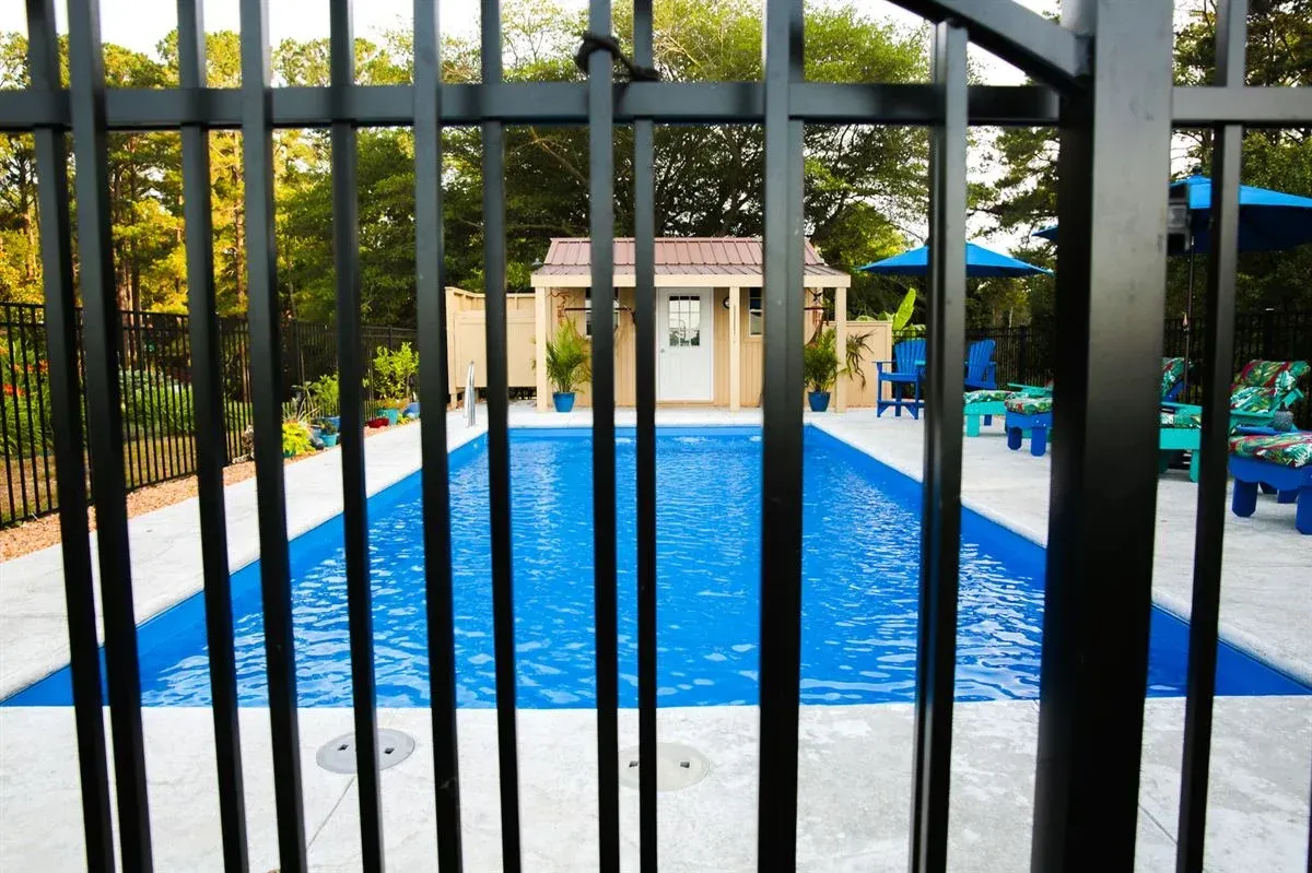 Black fence bars in front of a blue swimming pool and patio area with a small building beyond