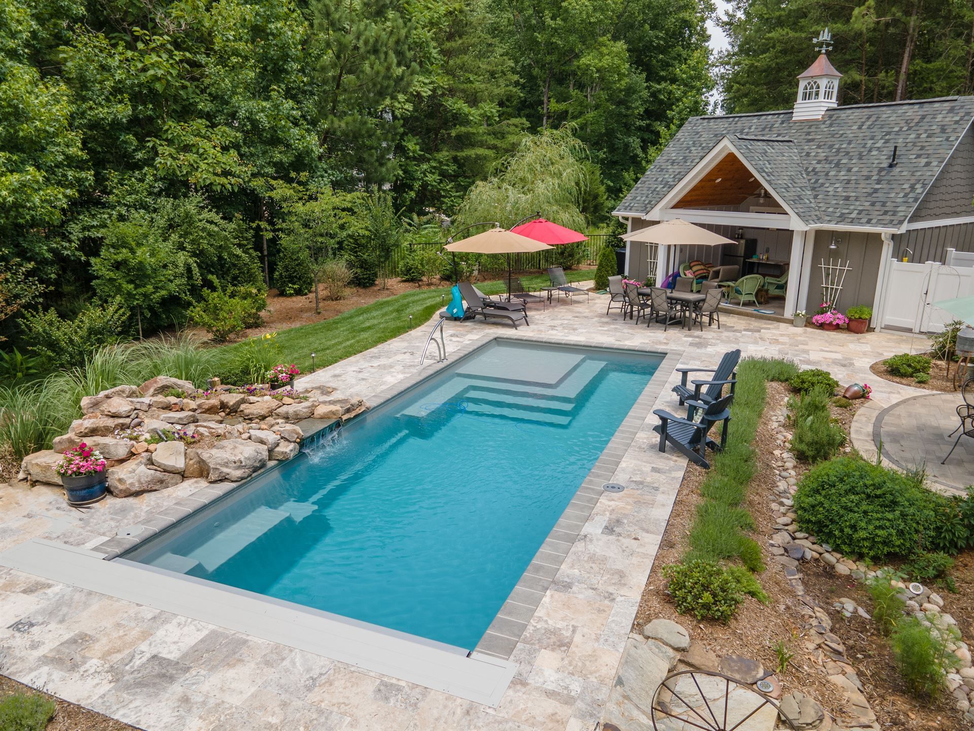 Backyard pool beside a stone patio and cottage, with lounge chairs and red umbrella in a lush garden
