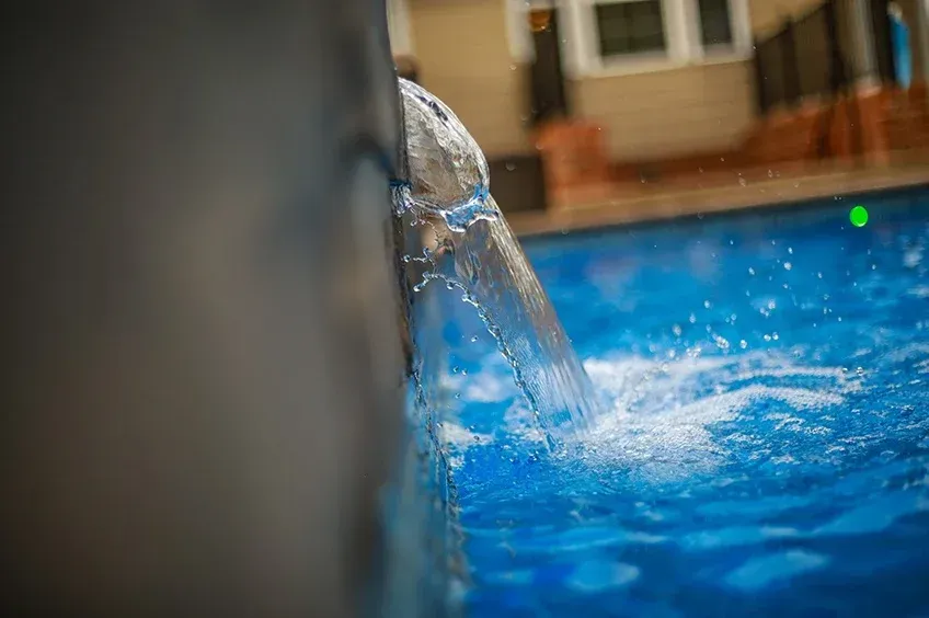 Pool water splashing from a pipe into a blue swimming pool near a house wall