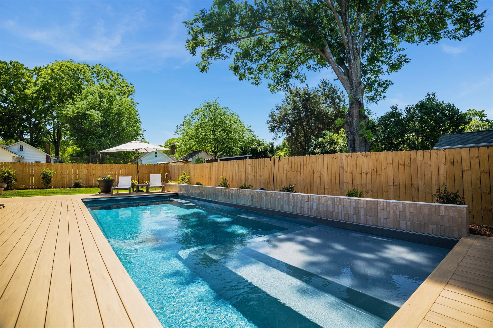 Backyard swimming pool with blue water, wooden deck, fence, and trees under a clear sky