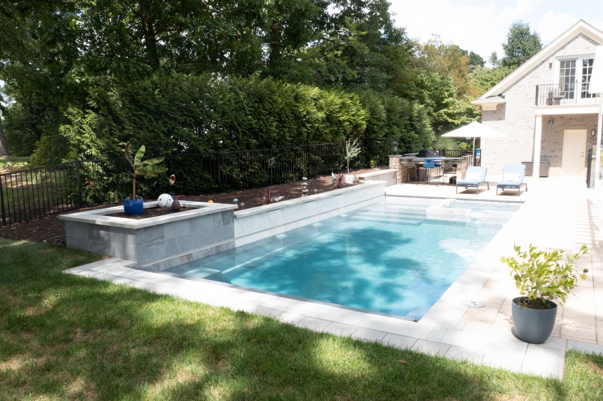 Backyard swimming pool with stone patio, lounge chairs, and lush green trees beside a house
