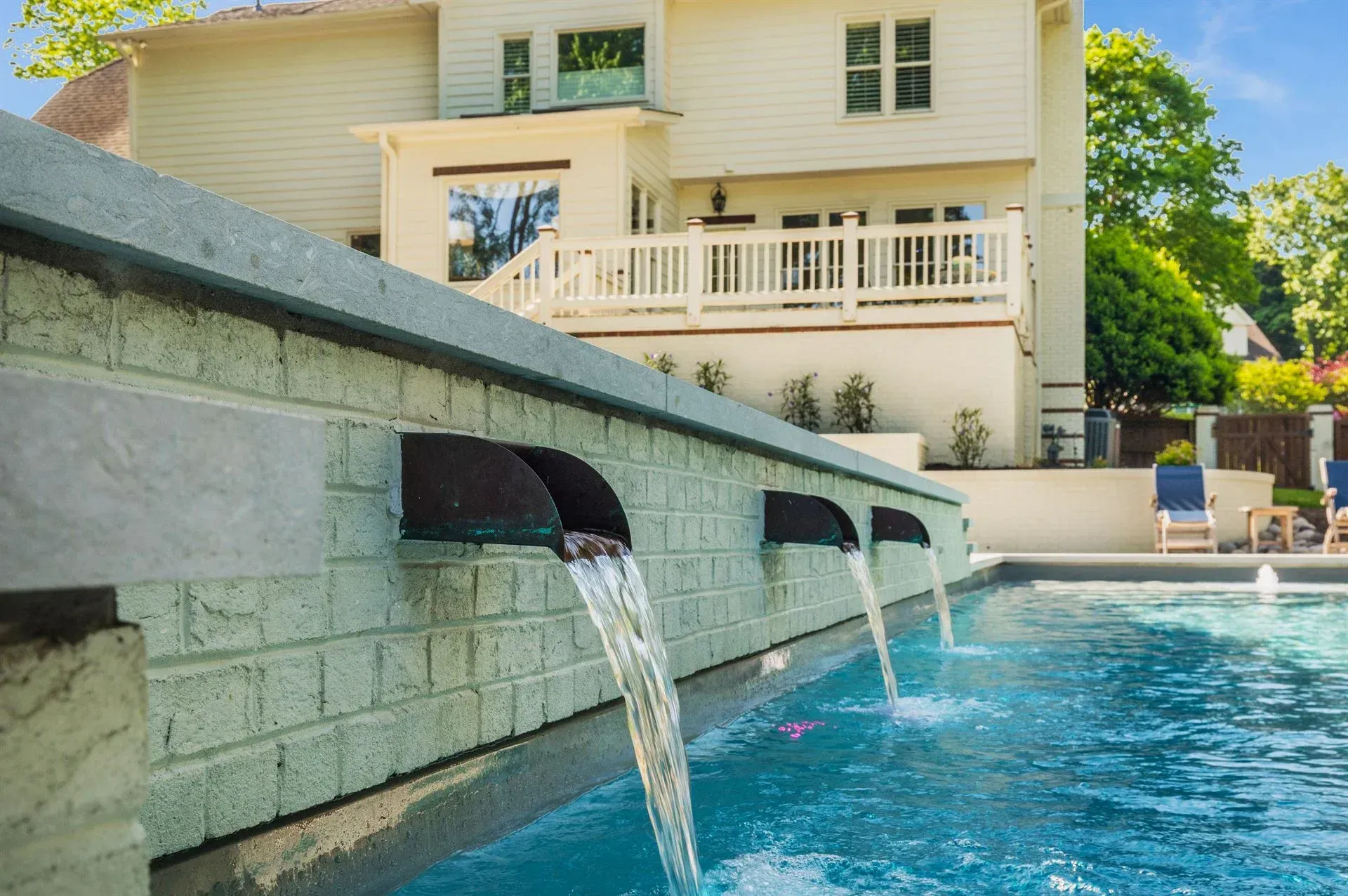 Outdoor pool with three wall waterfalls beside a cream-colored house and patio chairs
