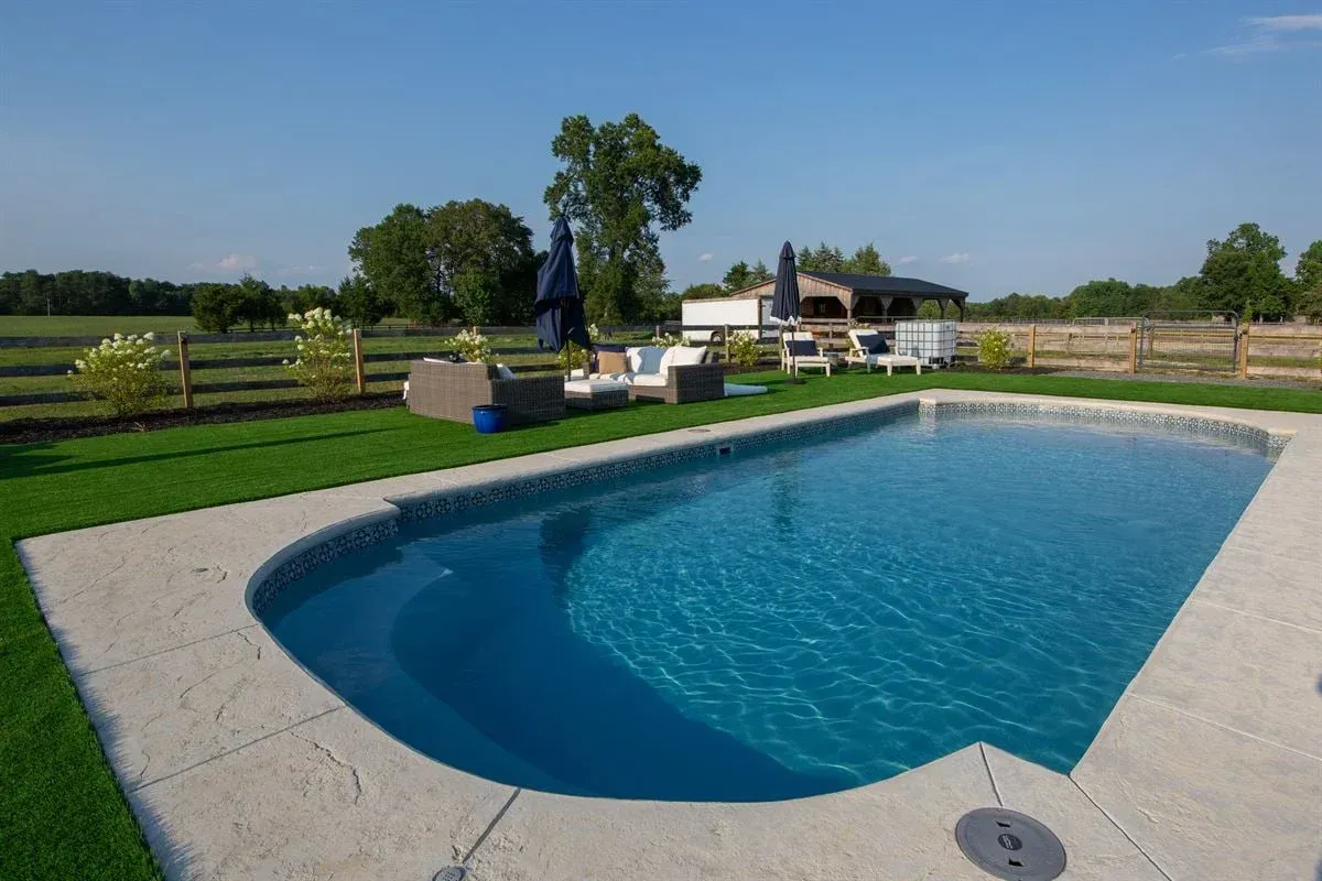 Backyard swimming pool with blue water, patio, lounge chairs, and a fenced lawn under a clear sky