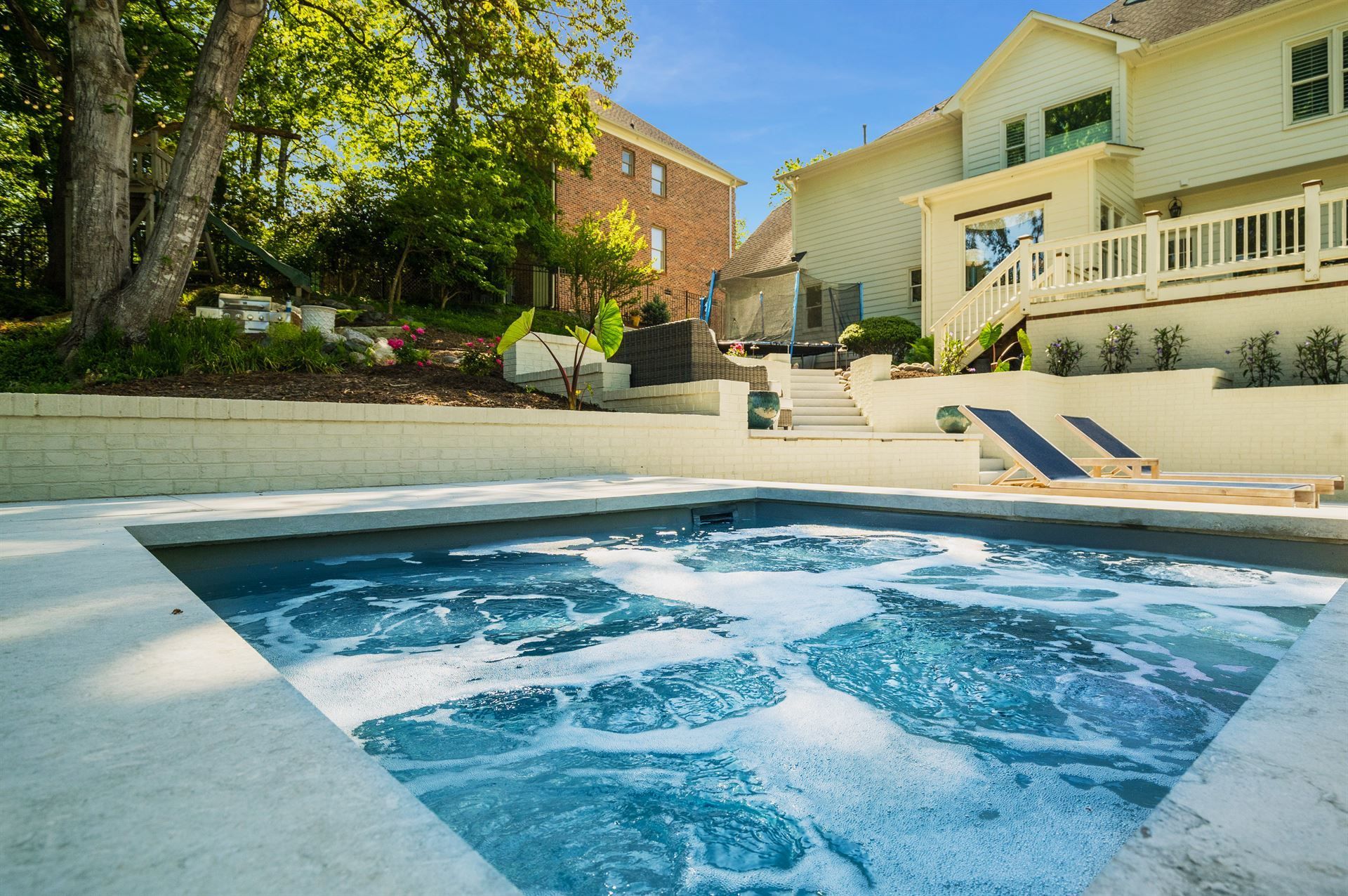 Backyard pool with bubbling water beside a sunlit house and stairs leading to a deck