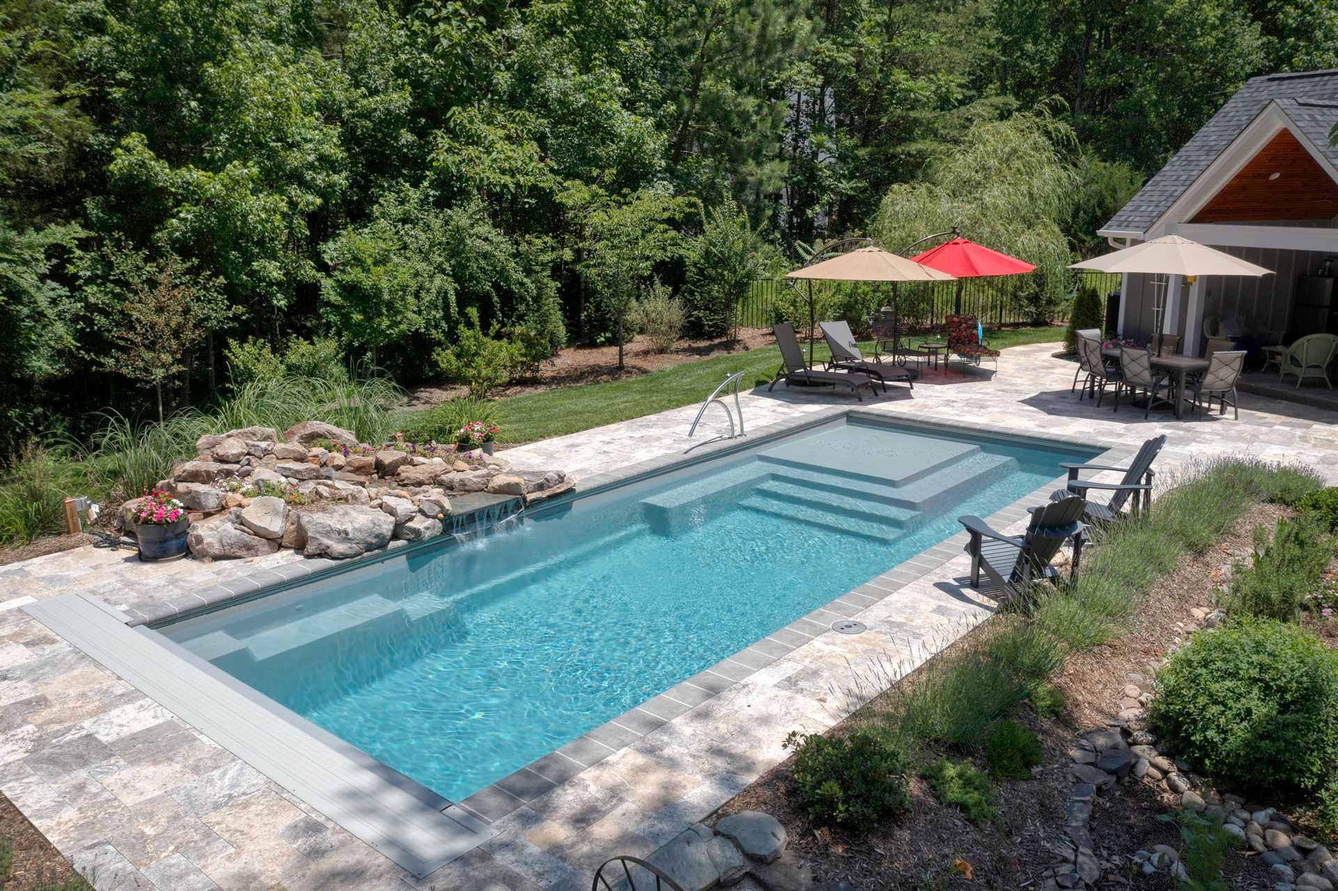 Backyard pool with stone patio, lounge chairs, red umbrella, and a wooded backdrop