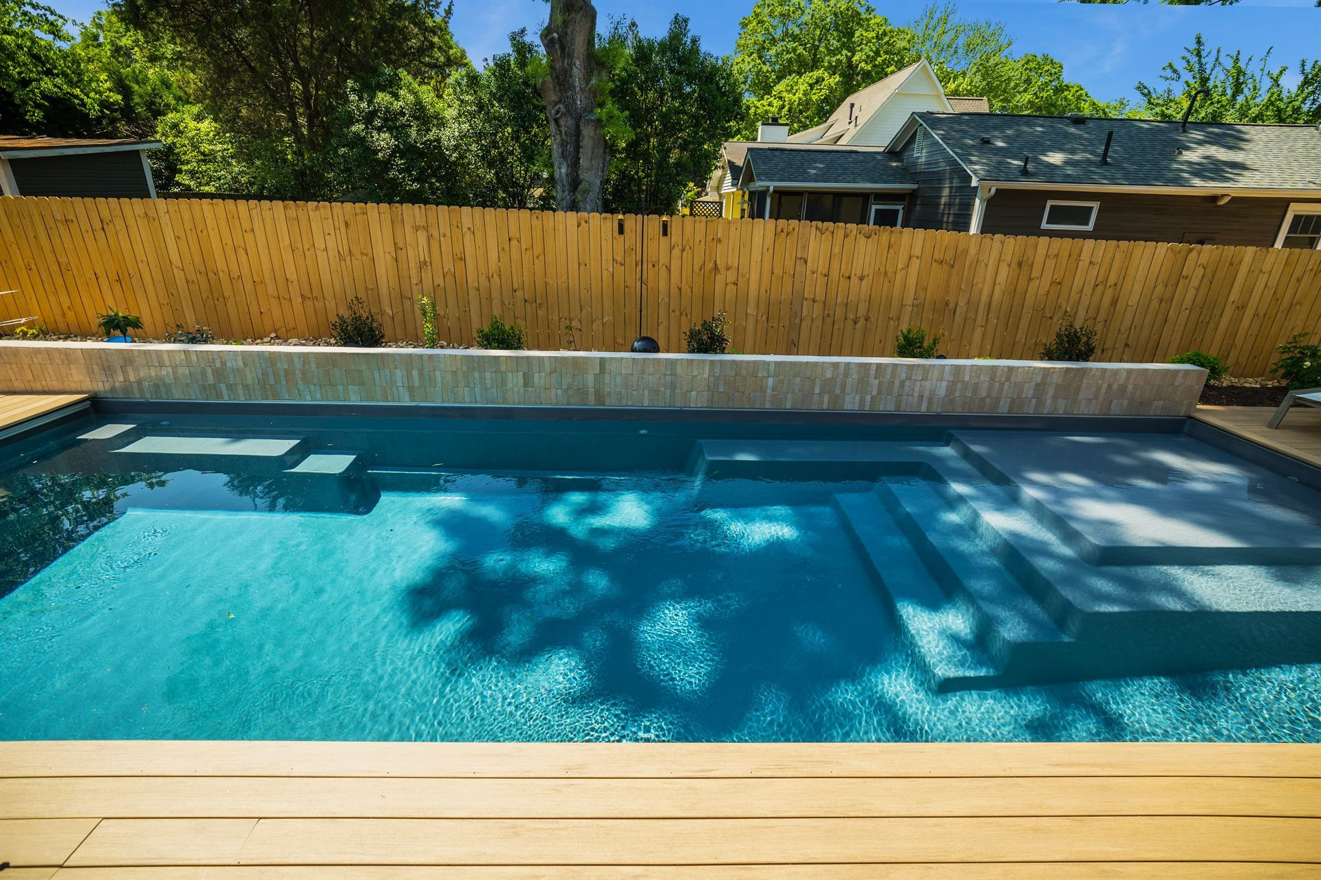 Backyard swimming pool with blue water, wooden deck, and fenced yard in sunlight