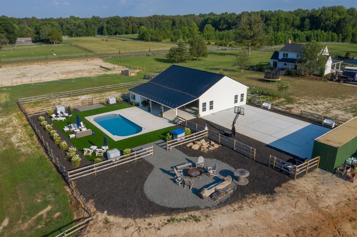 Aerial view of a modern white house with black roof, pool, patio seating, and fenced yard in a rural setting