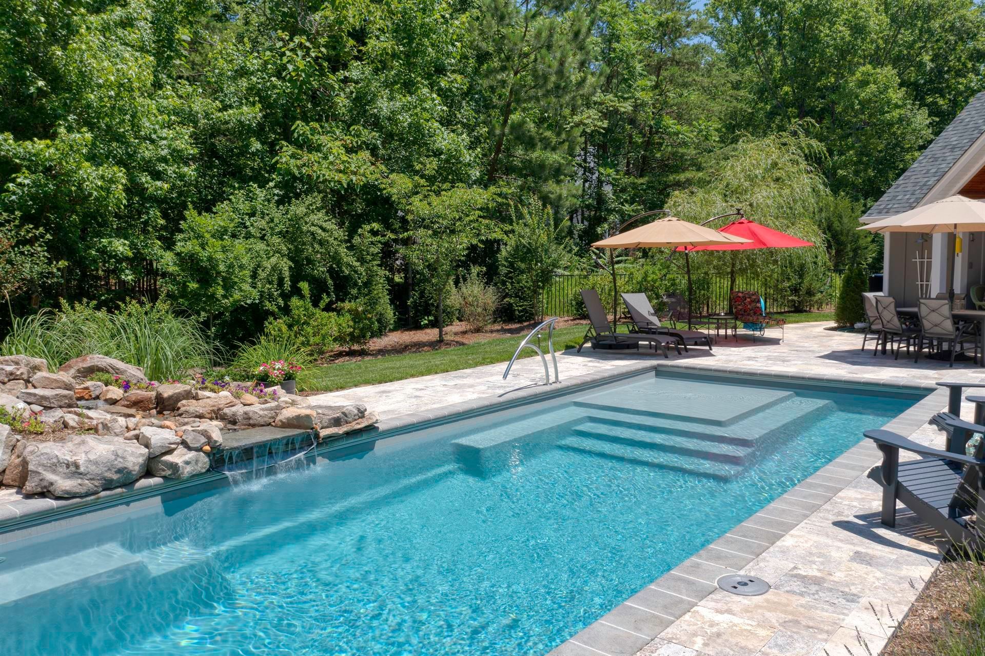 Backyard pool with turquoise water, stone patio, lounge chairs, and a red umbrella beside trees