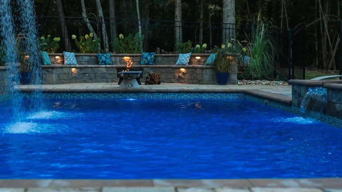 Blue-lit backyard swimming pool at night with a stone deck and lounge chairs along the far side