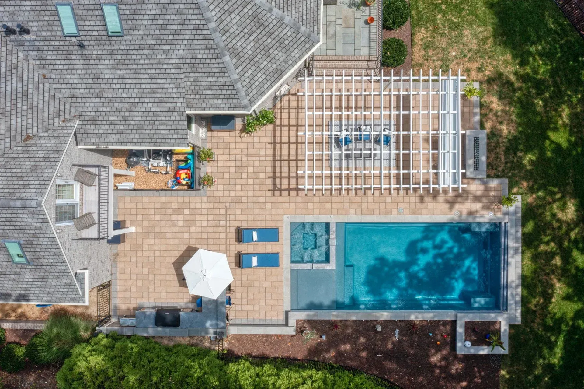Aerial view of a backyard patio with pool, lounge chairs, pergola, and a house roof beside greenery