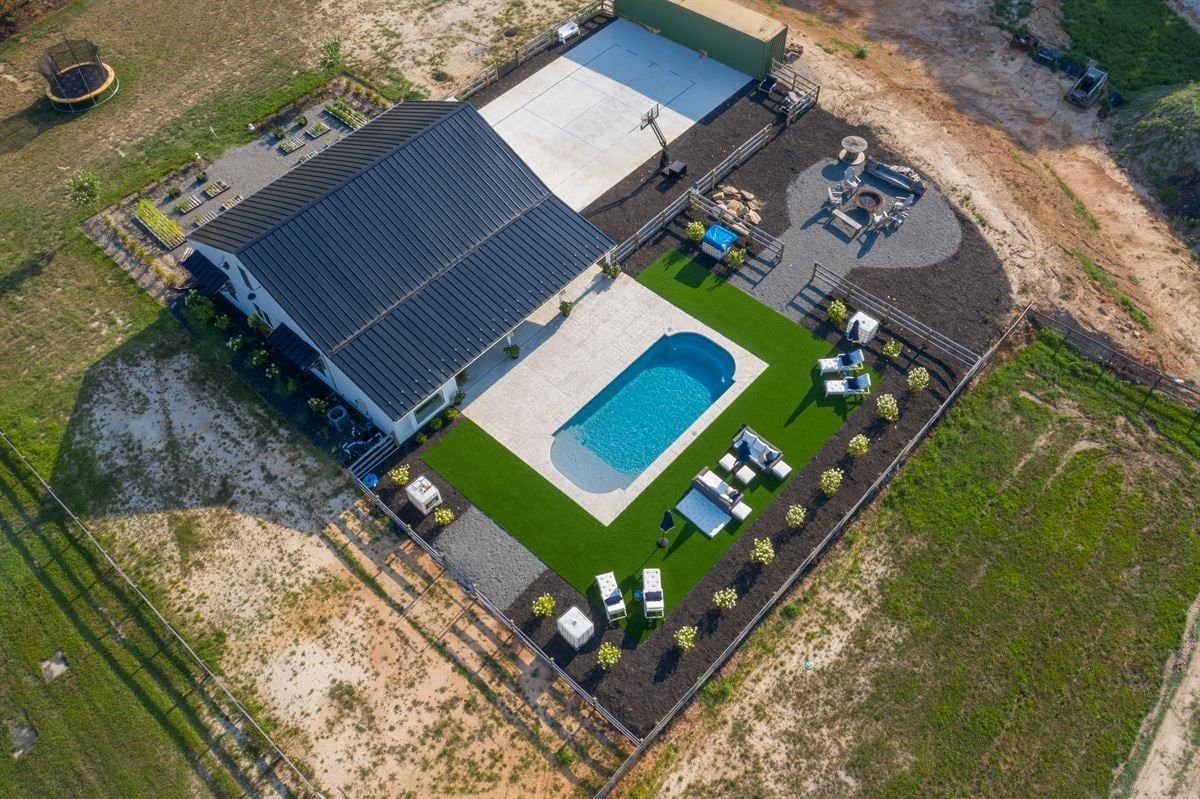 Aerial view of a house with a backyard pool, green lawn, and patio furniture in a rural setting