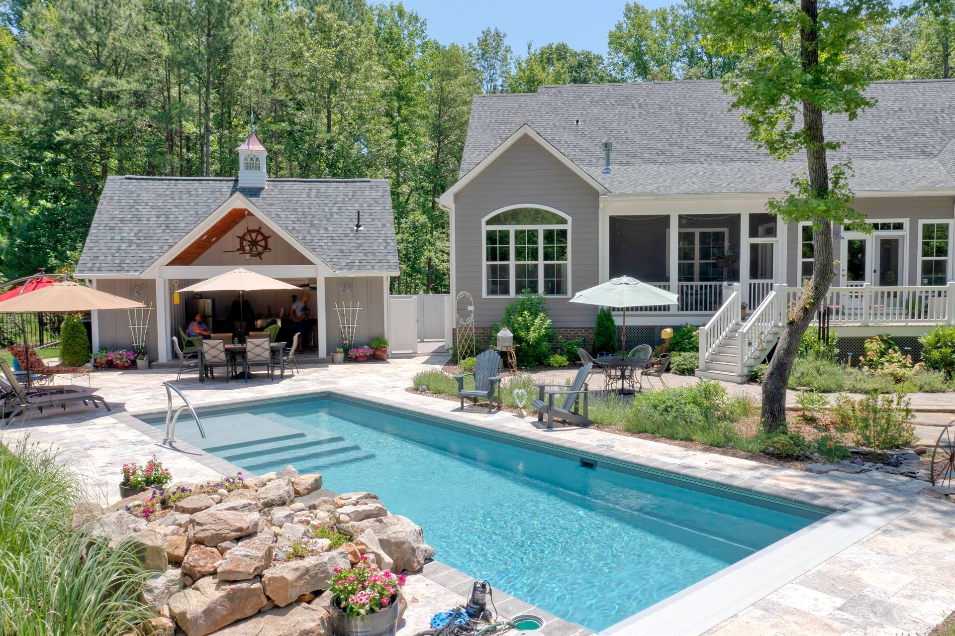 Backyard pool beside a gray house and patio with outdoor seating, surrounded by stone landscaping and trees