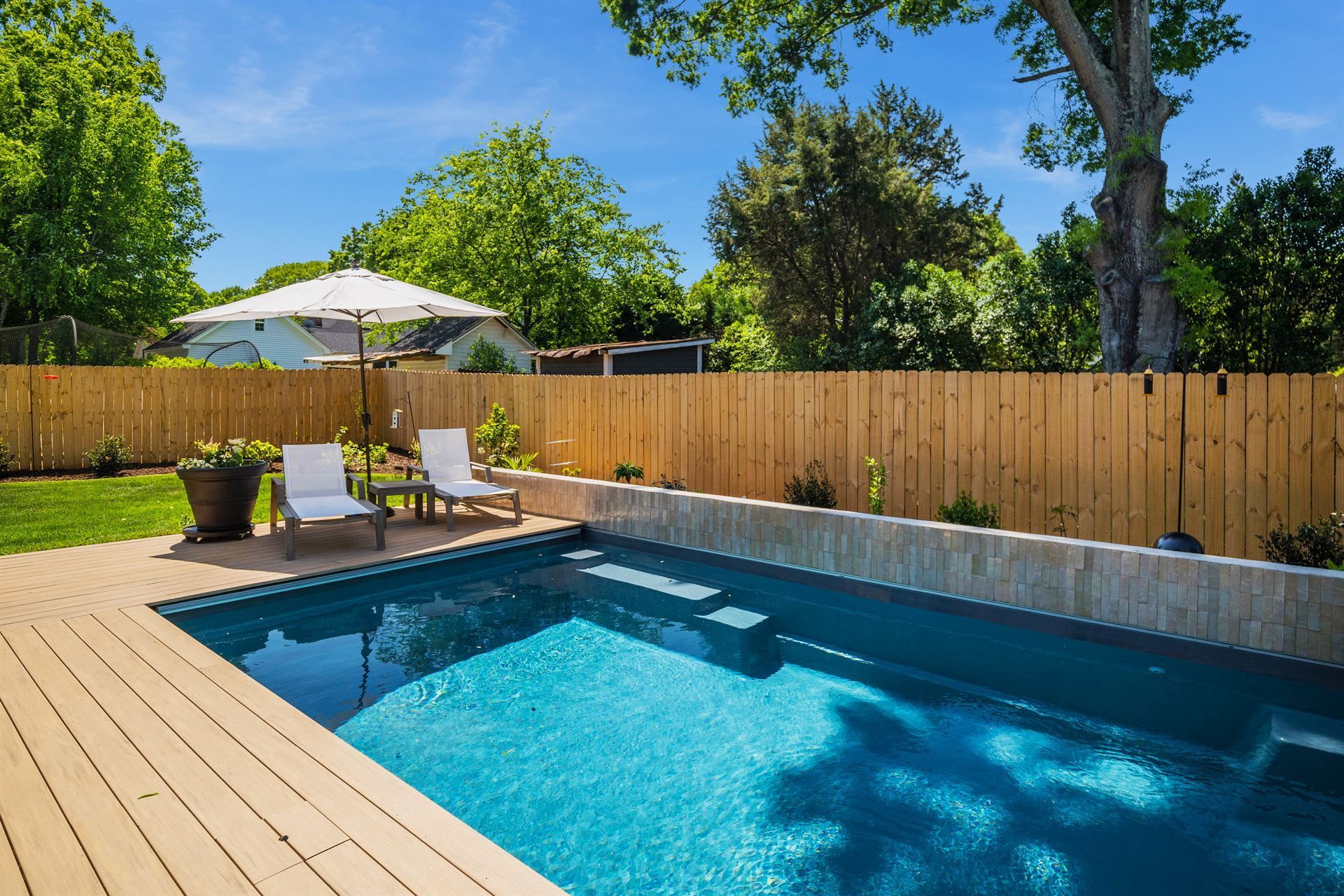 Backyard pool with wooden deck, lounge chairs, and umbrella beside a fenced yard and trees