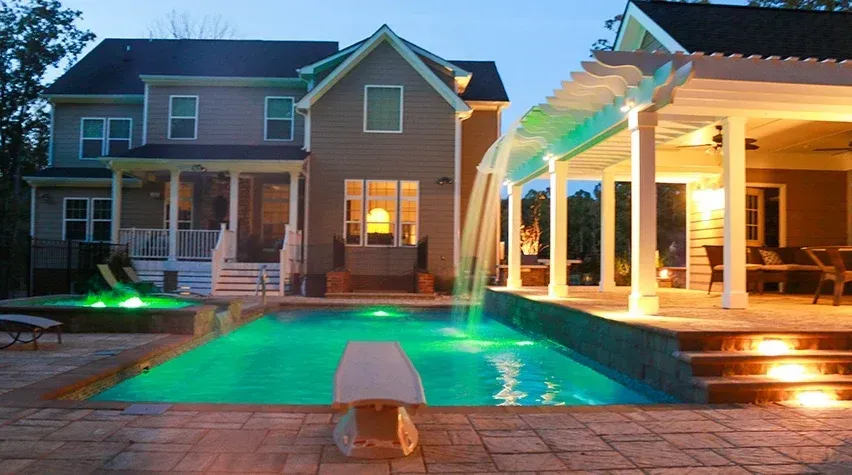 Backyard house with illuminated pool, deck lights, and covered patio at dusk