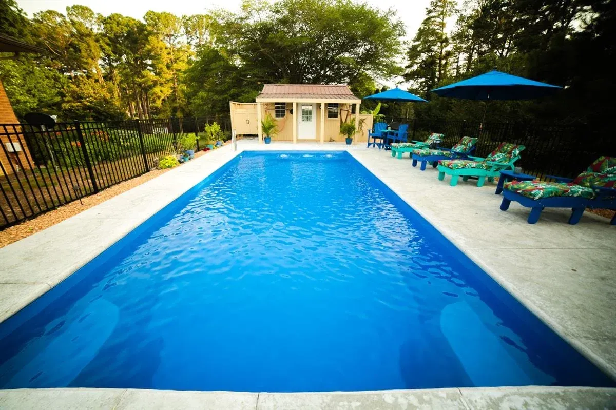 Outdoor swimming pool with bright blue water, lounge chairs, umbrellas, and trees surrounding it