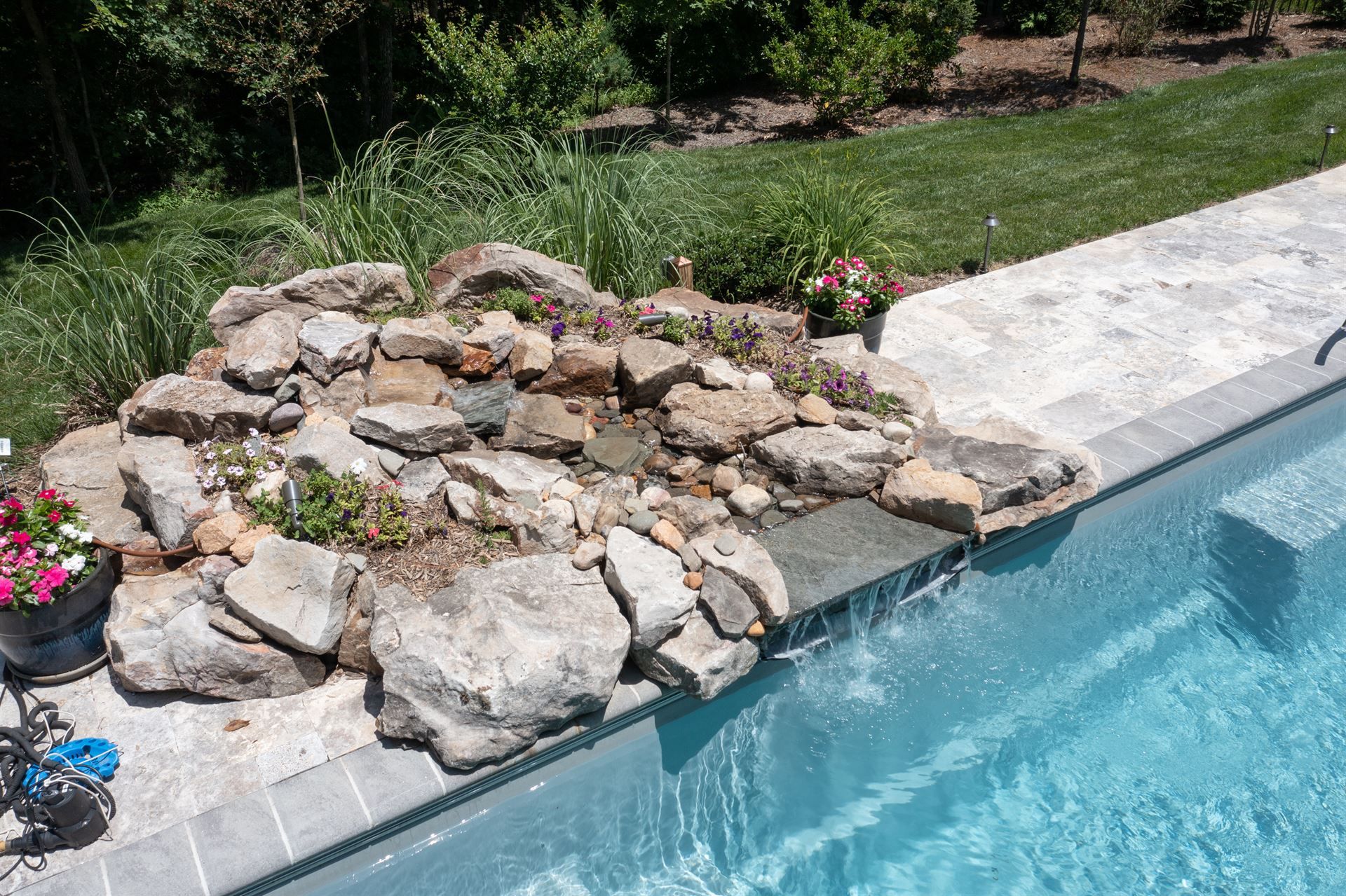 Rock waterfall feature beside a swimming pool and stone patio in a sunny backyard