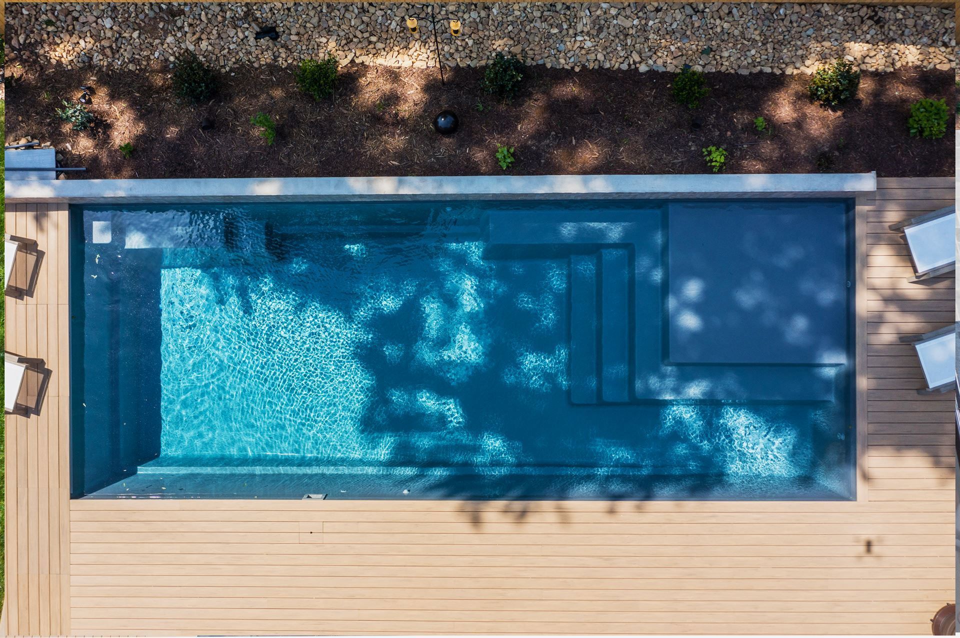 Top-down view of a rectangular blue swimming pool beside a wooden deck and stone garden border.