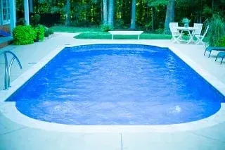 Blue backyard swimming pool surrounded by a patio, lounge chairs, and trees
