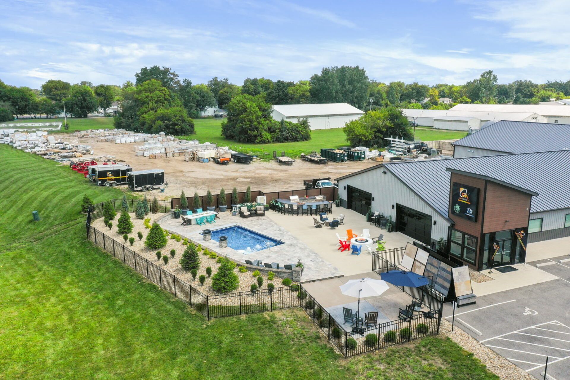Aerial view of an outdoor pool and patio beside a modern building, with tables, umbrellas, and grassy grounds.