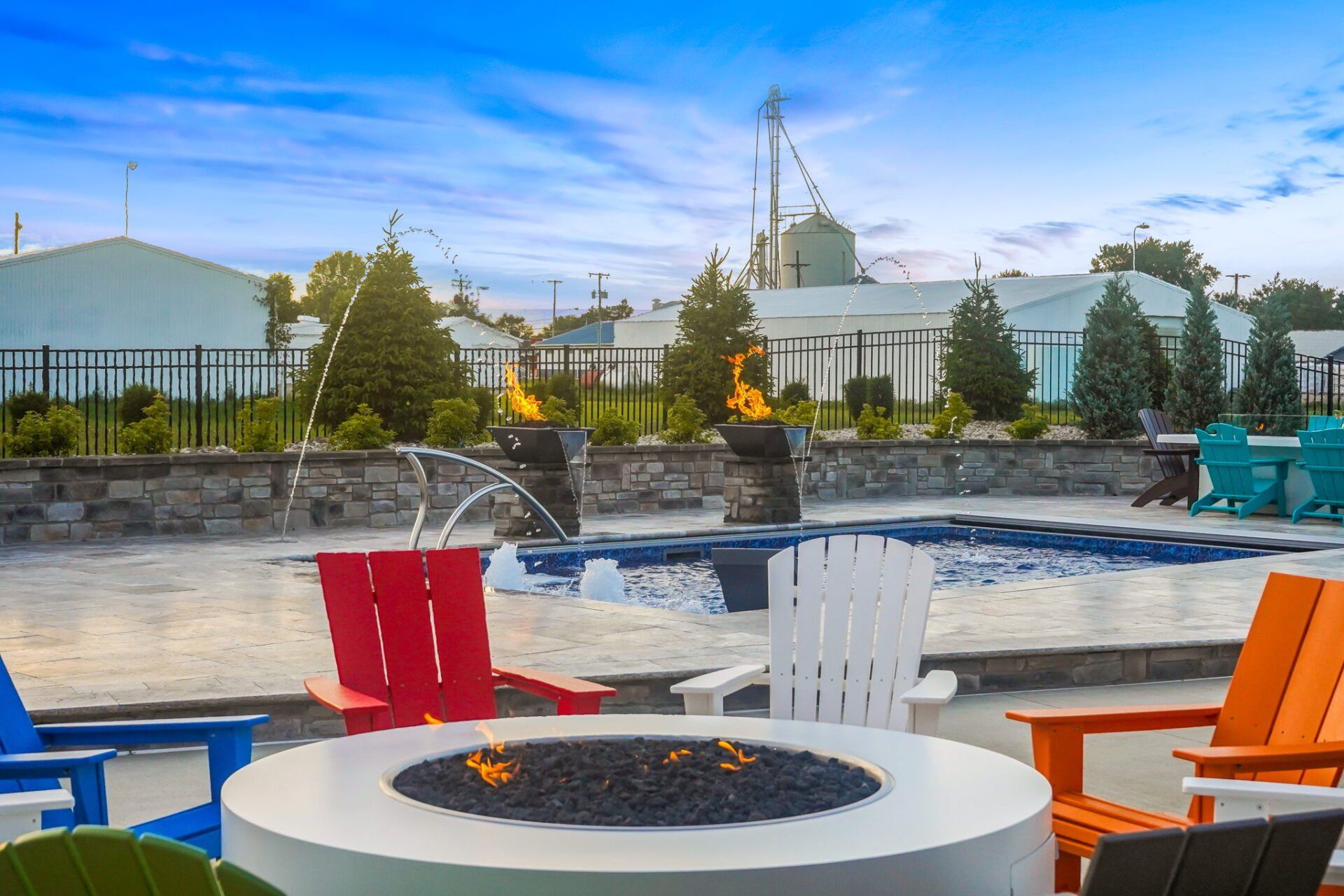 Outdoor patio with colorful chairs around a fire pit beside a pool and modern buildings beyond